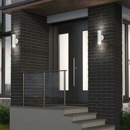 Modern front entrance with dark brick walls, glass door panels, metal railing, and two Artika C7 indoor-outdoor LED wall lights (3 CCT) on either side of the door. Concrete steps lead up to the entryway.