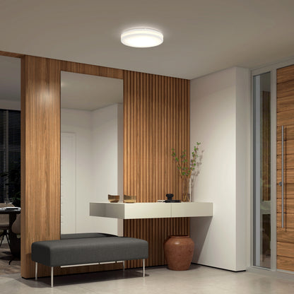 A modern entryway features wood panel walls, a large mirror, a floating white console with decor, a gray cushioned bench, a large vase, and an Artika Hemisphere 15 in. LED flush mount light. Glass doors and windows appear on the right.