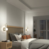 A modern bedroom with a rattan headboard, gray and white bedding, rust-colored accent pillows, two wall sconces, a wooden nightstand, and a window with white blinds letting in soft natural light.