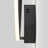 A modern black wall-mounted light fixture with a curved LED bar, emitting a soft, warm glow. The fixture is attached to a pale wall and features a visible screw on its rectangular mounting plate.