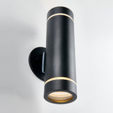 A modern black cylindrical wall sconce with thin gold accents, mounted vertically on a white wall, emitting light from both the top and bottom ends.
