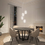 A modern dining room with four beige upholstered chairs around a round black table, hanging pendant lights above, a fireplace with a lit fire, large window with sheer curtains, and a potted plant in the corner.