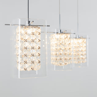 The Artika Crystal Cube 3-light LED pendant light features three modern fixtures with clear glass and geometric crystal-like patterns, hanging in a row and casting a warm, bright glow against a light gray background.