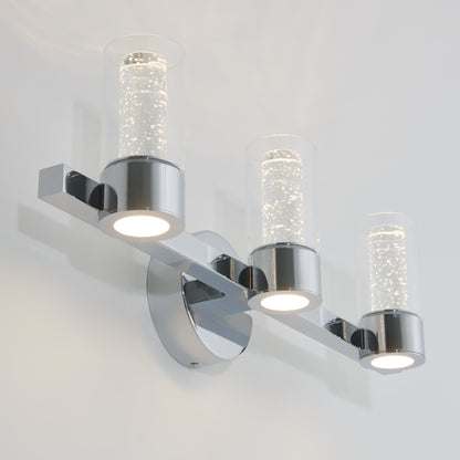 The Artika Novello modern LED bathroom vanity light features three cylindrical glass shades with bubble details, mounted on a chrome base against a white wall.