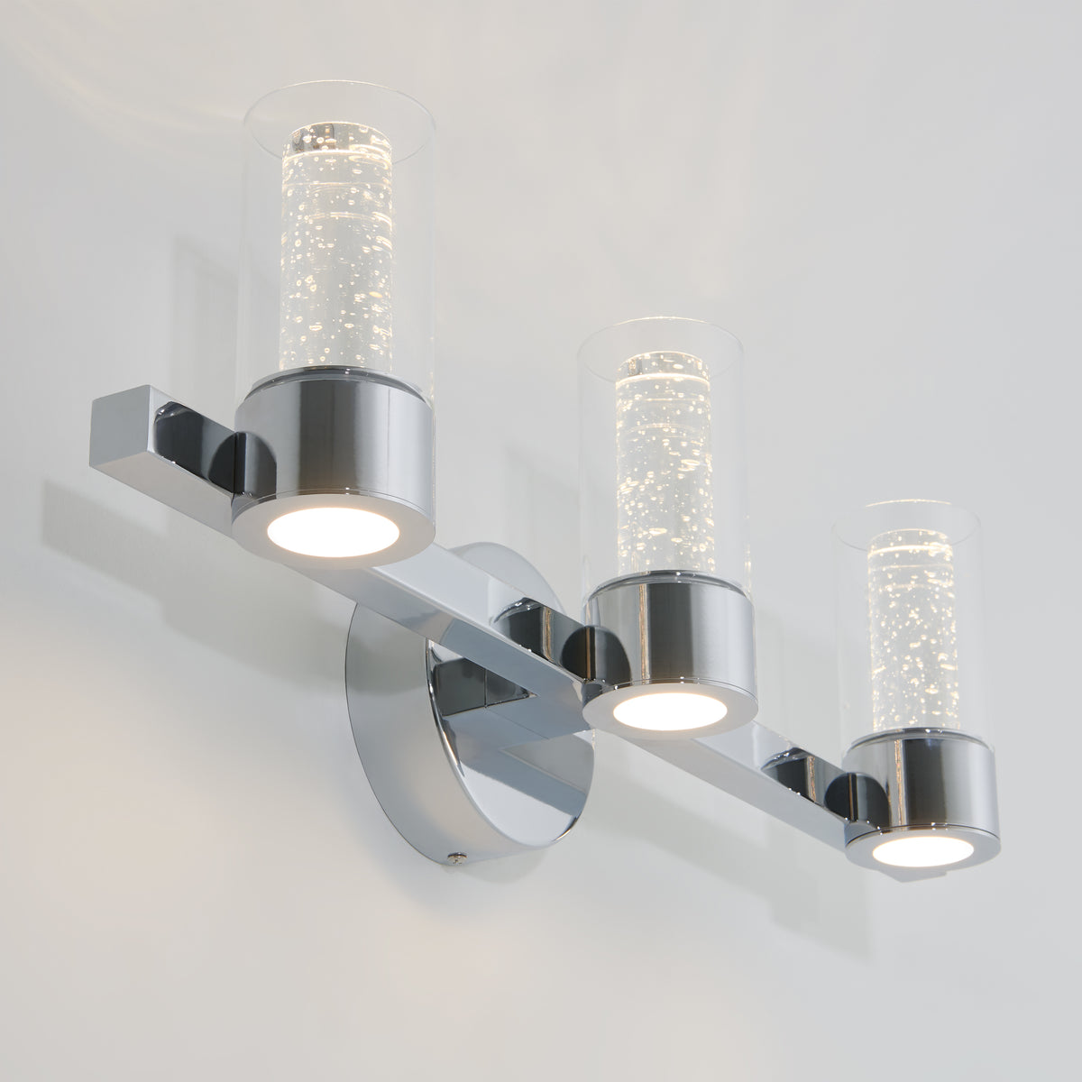 A modern wall-mounted light fixture with three cylindrical glass shades, each containing bubbles, mounted on a chrome base and arm, emitting a soft white light.