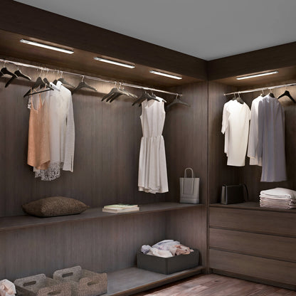 A modern walk-in closet with dark wood shelves, white and beige clothing, baskets, towels, a pillow, a shopping bag, and soft overhead lighting from the Artika Outline under cabinet LED light 3CCT.