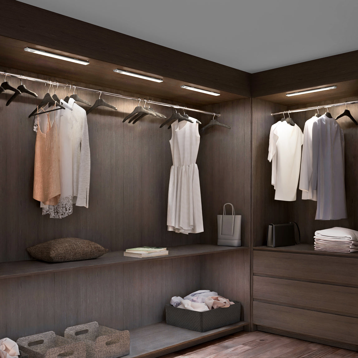 A modern walk-in closet with wooden shelves and hanging rods holding white and beige clothes on hangers, folded garments, storage baskets, a pillow, and a shopping bag, all under built-in overhead lighting.