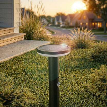An Artika Urbia Solar LED Bollard with Motion Sensor stands in grass by a porch, houses and sunset visible behind.