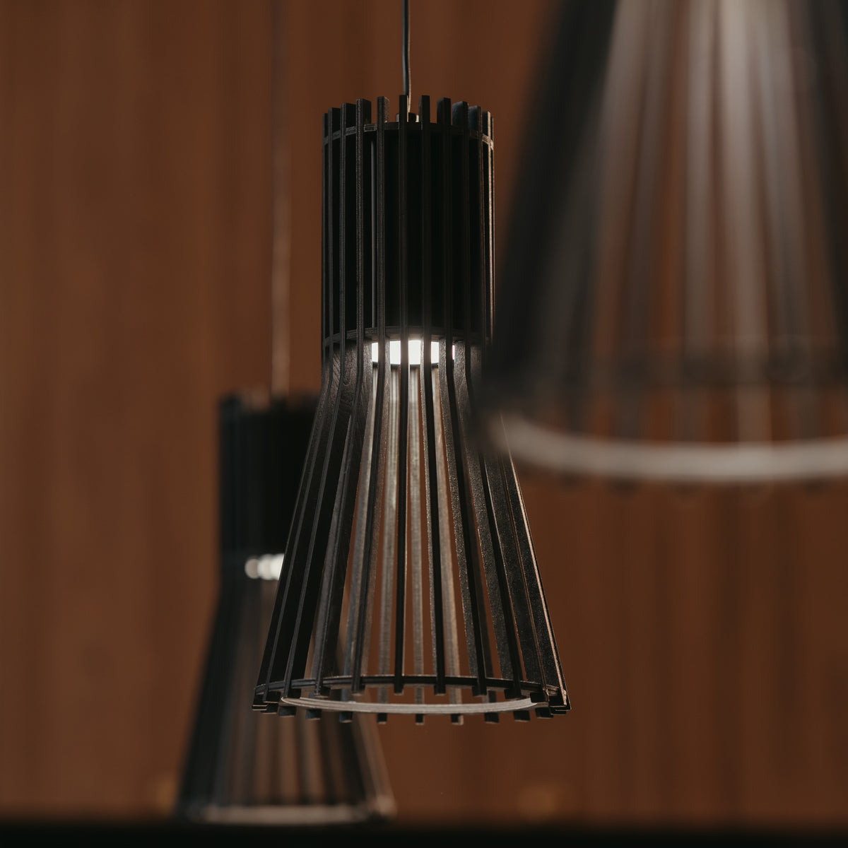 Close-up of modern, black pendant lights with vertical slats, hanging from the ceiling. The background is blurred and warm-toned, highlighting the sleek design of the lighting fixtures.