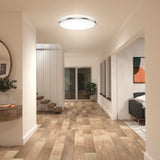 A modern hallway with wood flooring, a round ceiling light, a round mirror above a console table with a plant, stairs with white railings, large windows, and a glimpse of a living area with a sofa and wall art.