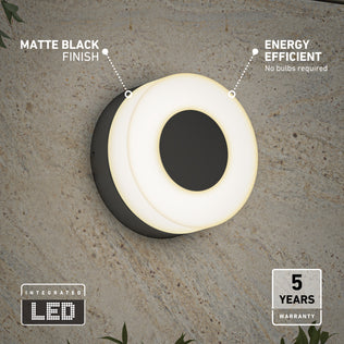 The Artika Ludwig modern LED indoor-outdoor wall light 5CCT features a matte black finish and glowing white LED ring. Energy efficient with integrated LEDs—no bulbs needed—and comes with a 5-year warranty. Shown mounted on a textured wall.