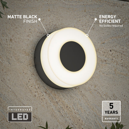 The Artika Ludwig modern LED indoor-outdoor wall light 5CCT features a matte black finish and glowing white LED ring. Energy efficient with integrated LEDs—no bulbs needed—and comes with a 5-year warranty. Shown mounted on a textured wall.