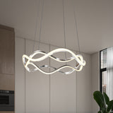A modern, sculptural LED pendant light with intertwined loops hangs from the ceiling in a contemporary room with light walls, wood cabinetry, a window, and a green plant.