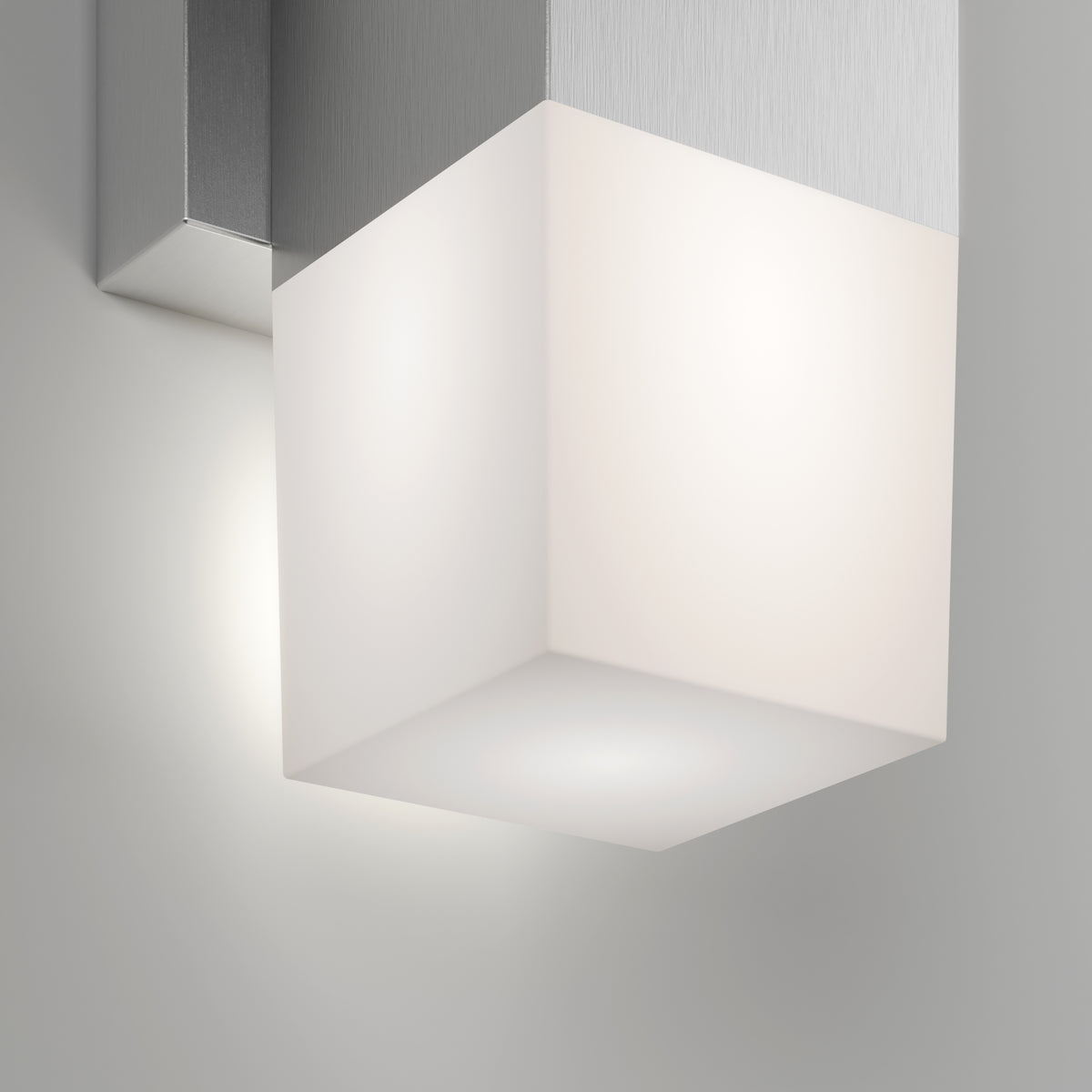 A modern wall-mounted light fixture with a frosted glass cube shade and a metallic base, softly illuminating the surrounding white wall.