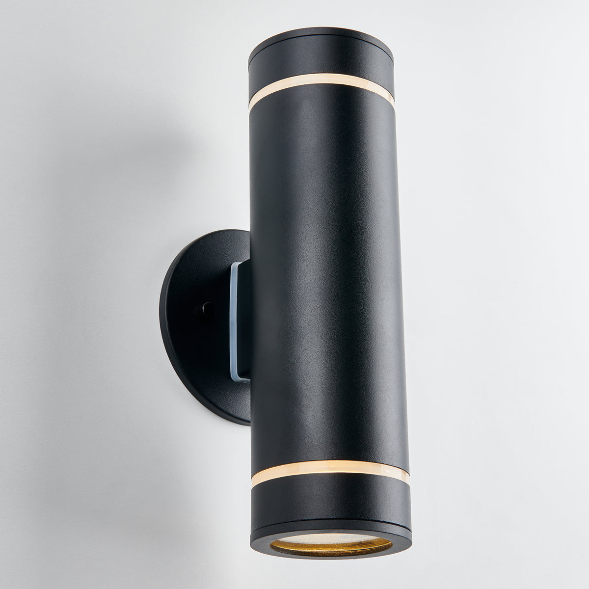 The Artika C7 indoor-outdoor wall light is a modern black cylindrical sconce with gold accents, rustproof, mounted vertically on a round base, and designed to cast light up and down for versatile lighting needs.