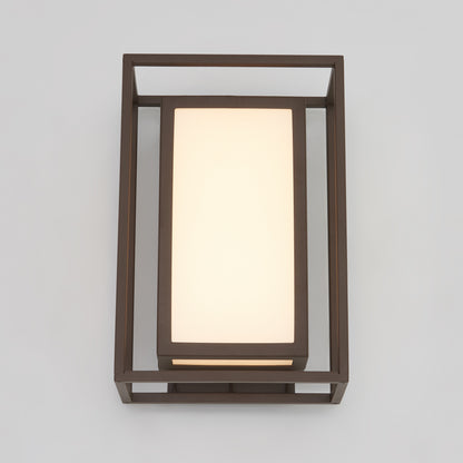 The Artika Ghost indoor-outdoor LED wall light features a rectangular frosted glass shade framed by a dark metal geometric structure, stylishly mounted on a light gray wall.