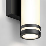 Close-up of a modern wall-mounted cylindrical light fixture with a matte black frame and frosted white glass, emitting a soft, even glow against a light-colored wall.