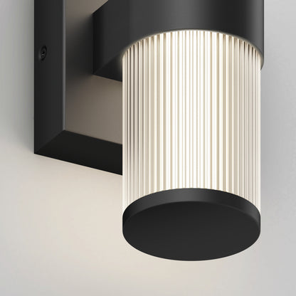 Close-up of the Artika Mirado modern LED wall sconce 5 CCT, featuring a ribbed translucent cylindrical shade and matte black base, casting a soft, diffused glow on a light-colored wall.