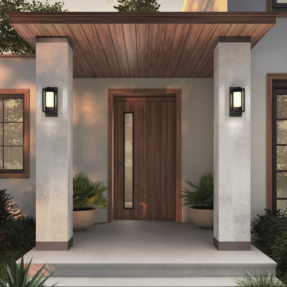 Modern front porch featuring a wooden door, vertical window panel, overhanging wooden ceiling, concrete pillars, potted plants flanking the entrance, and Artika’s Aurel Outdoor LED Light (3CCT) for sleek black rectangular wall lighting.