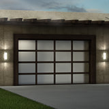 A modern garage door with frosted glass panels and a dark frame, flanked by two illuminated wall-mounted lights on a stone exterior, adjacent to a concrete driveway and a patch of grass.