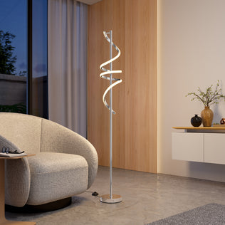 A modern living room featuring a textured gray armchair, the Artika Coda modern LED floor lamp 5CCT with a twisted design, a wooden accent wall, floating sideboard, and a vase with branches by a large window with sheer curtains.