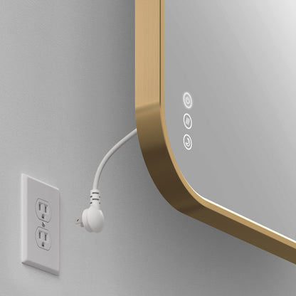 An Artika Royce Dimmable LED Mirror (3 CCT, mountable horizontally or vertically) is reflected in a gold frame; a white cord plugs into a white outlet on a gray wall, illuminating its adjustable LEDs and touch buttons.