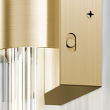 Close-up of a modern gold wall sconce with a brushed metal finish, featuring a circular button and a screw. Clear vertical glass rods hang below, reflecting light. The background is a plain white wall.