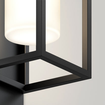 Close-up of the Artika Aurel Outdoor LED Light, 3CCT— a modern wall-mounted fixture with a black metal geometric frame and frosted glass cylinder shade, casting a soft white glow on a light-colored wall.