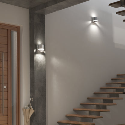Modern interior with floating wooden stairs, two Artika Valor Round LED wall lights casting upward and downward beams, a wooden door with vertical handle, umbrella stand with umbrellas, and neutral-toned walls.