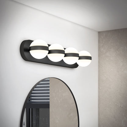 The Artika Baldric LED modern bathroom vanity light 3 CCT, featuring four spherical frosted glass bulbs on a black horizontal base, is mounted above a round mirror on a white wall reflecting a window with blinds.
