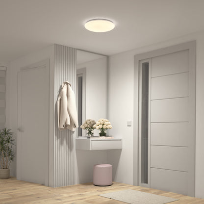 A modern hallway features light wood flooring, white walls, an Artika Empress 13 in. LED modern flush mount light, a small floating shelf with flowers and decor, a pink stool, and a beige coat hanging on a wall panel.