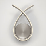 A modern wall-mounted light fixture with two curved metallic arms crossing at the top, forming a teardrop shape around a circular, brushed metal base. The fixture has LED strips on the inner edges of the arms.