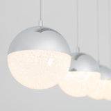 Three modern spherical pendant lights with metallic tops and textured, glowing bottoms are hanging in a row against a plain, light background. The focus is on the nearest light, with the others blurred behind it.