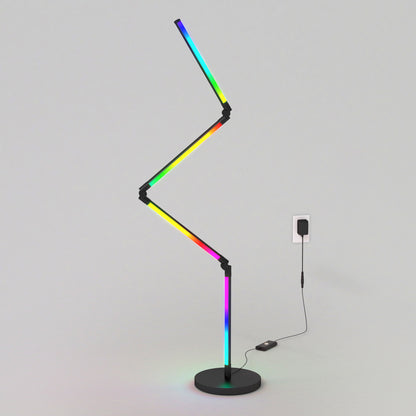 The Artika Fitch smart foldable RGB LED gaming floor lamp, featuring a modern zigzag shape and black base, displays multiple colors while plugged into a wall outlet against a plain light-gray background.