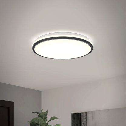 The Artika Macy 15 in. LED modern flush mount light (RGB Wifi) with a black rim is lit on a white ceiling. A green plant and wall art appear in the lower corner of the image.
