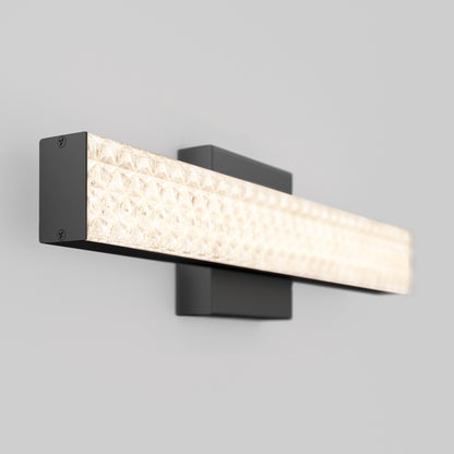 The Adeline modern LED bathroom vanity light by Artika features a rectangular black metal base and a long, textured illuminated glass bar, mounted on a plain light gray wall. Three selectable color temperatures (CCT) are included.
