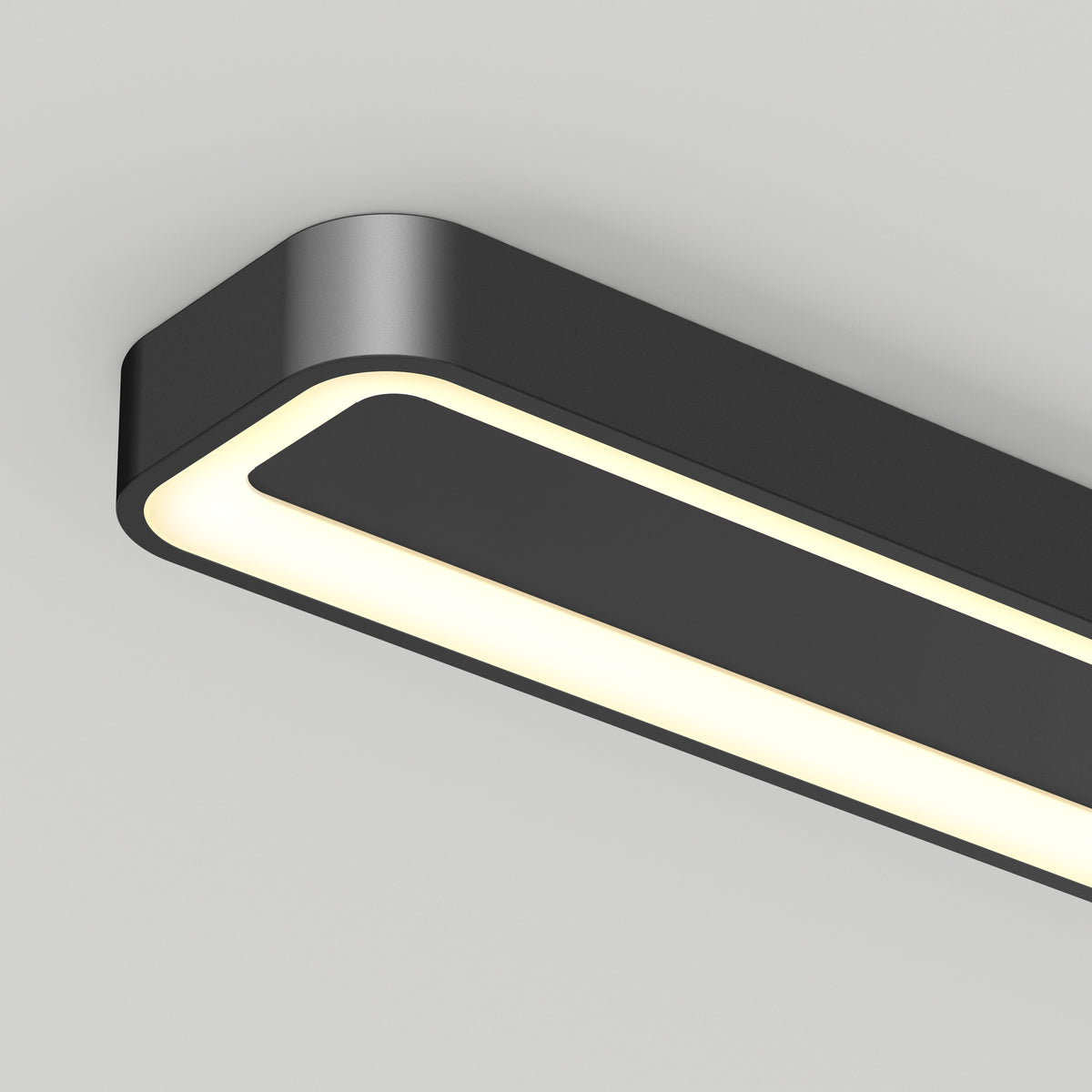 A close-up view of a modern, rectangular black ceiling light fixture with rounded edges, emitting a soft white glow. The fixture is mounted on a plain white ceiling.