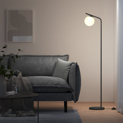 A modern living room features a gray sofa, light gray pillow, geometric wall art, coffee table with a glass, a plant, and the Artika Clayton dimmable LED modern mid-century floor lamp with a glowing round bulb. Light filters through sheer curtains.