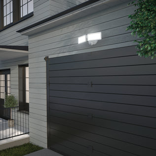 The Artika Sentinel outdoor Flat Panel Security light, a modern weatherproof spotlight with two bright panels, is mounted above a dark gray garage door on a light house, illuminating the area at dusk with greenery visible to the right.