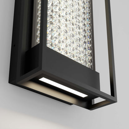 A close-up of the Artika Glam indoor-outdoor LED wall light features a black metal frame, clear crystal accents, and a bright LED shining downward against a white wall.