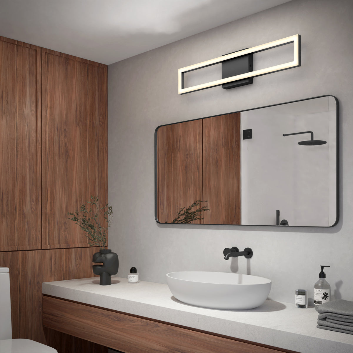 Modern bathroom with wooden cabinets, a large rectangular mirror, a white vessel sink on a light countertop, matte black fixtures, a geometric light fixture above the mirror, and minimalist decor.