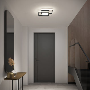 A modern hallway features an Artika Romeson 11 in. LED flush mount light, a dark door, a console with a vase of beige flowers on the left, large mirror above, and glass-railed stairs on the right.
