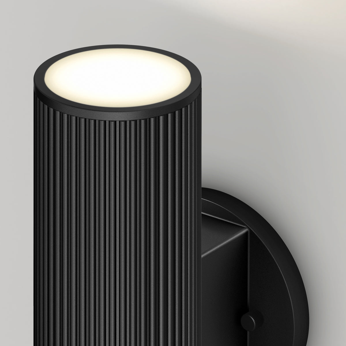 A close-up of the Artika Groove LED indoor-outdoor wall light, featuring a modern black ribbed cylindrical design and emitting soft white light from the top, mounted on a light-colored wall.