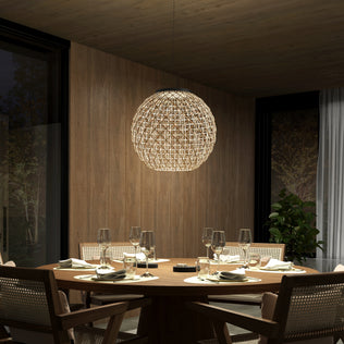 A modern dining room features a round wooden table for four, light-cushioned chairs, and the Artika Benson LED pendant light 5 CCT overhead. The lamp casts a warm glow over wood-paneled walls and a nearby plant.