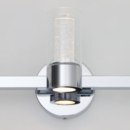 The Artika Novello modern LED bathroom vanity light features a chrome finish, a bubble-filled cylindrical glass shade, a circular base, and emits downward light.