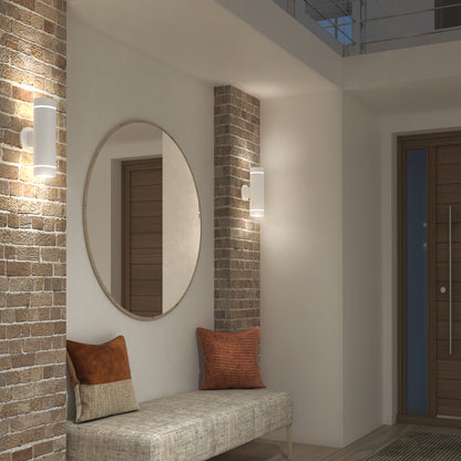 A modern entryway with exposed brick columns, a large round mirror, a cushioned bench with two accent pillows, an Artika C7 indoor-outdoor LED wall light 3 CCT, and a wooden front door with a vertical metal handle.