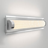A modern wall-mounted light fixture with a long, cylindrical, textured glass shade emits a soft, warm glow. The fixture features chrome accents and is mounted on a rectangular metal backplate.