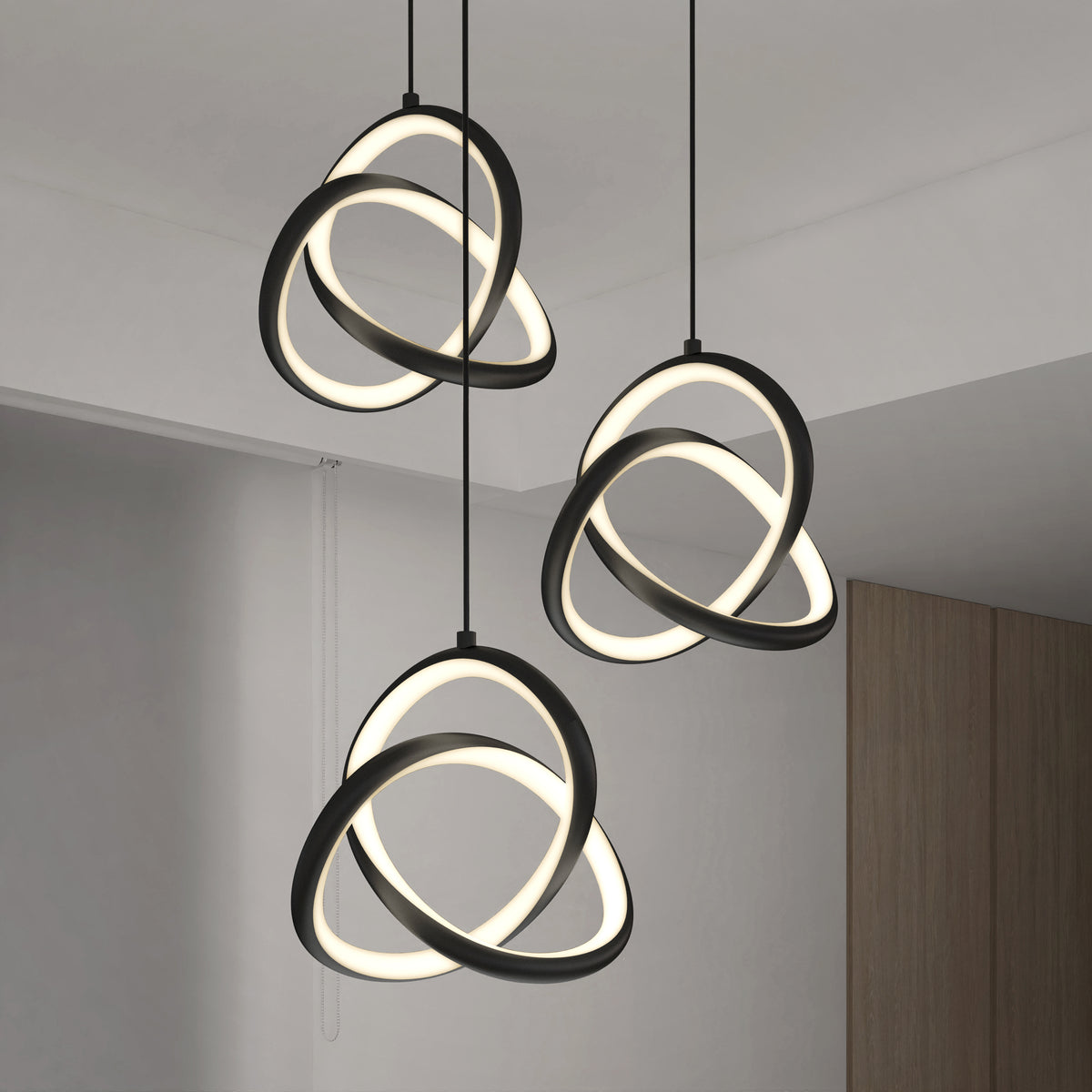 The Artika Zephyr 3-light LED pendant features three intertwined glowing rings suspended from the ceiling, adding a modern touch to a minimalist room with white walls and a wooden accent.