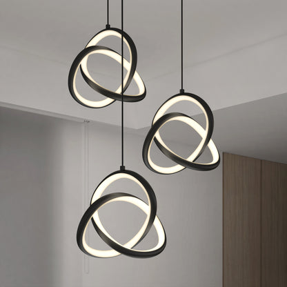 The Artika Zephyr 3-light LED pendant features three intertwined glowing rings suspended from the ceiling, adding a modern touch to a minimalist room with white walls and a wooden accent.