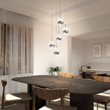 Modern dining room with a dark wooden table, four wooden chairs, pendant globe lights, sheer curtains on a window, and minimalistic wall art in a neutral-colored space. A sofa and coffee table are visible in the background.